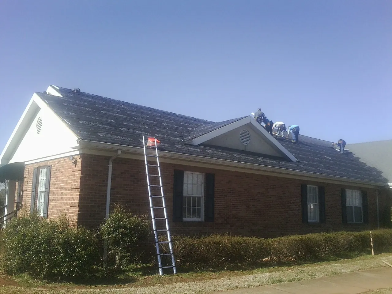 Crew tearing shingles off a brick ranch roof with a ladder set — Emergency Roof Repair work by our crew in Marrero