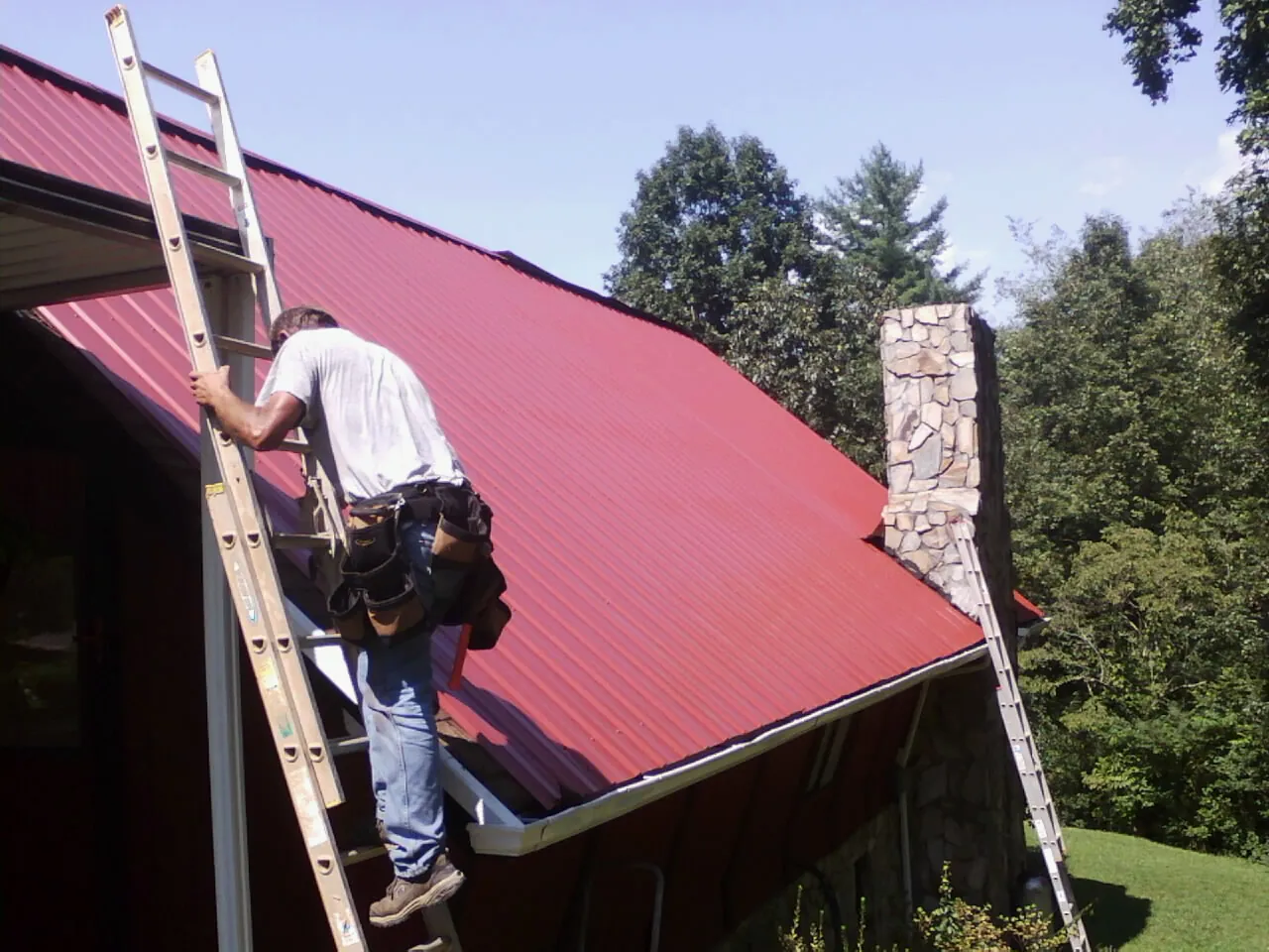 Roofer on a ladder beside a red metal roof and stone chimney — Soffit & Fascia Repair by Roofing Brothers in Marrero